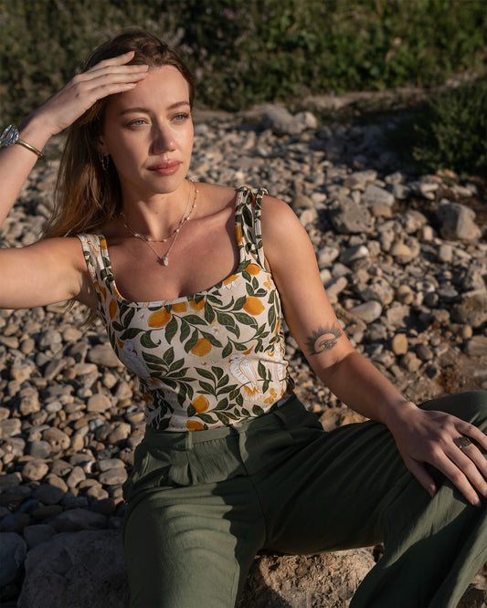Woman sitting at the beach wearing a patterned top and green pants. The top features a print of lemons, leaves, and cockatoos. 