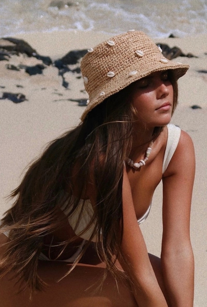 A tan crochet bucket hat with cowry shells woven on it for women's casual beachy style. 