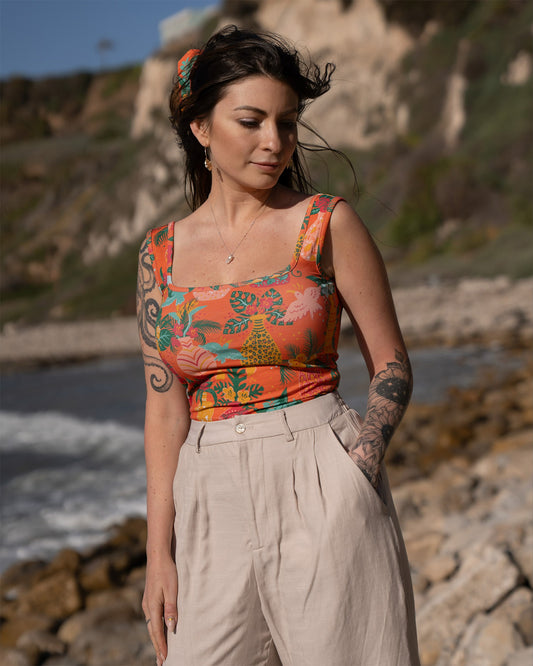 Woman wearing a colorful sleeveless top with a tropical floral print and beige pants standing on a rocky beach.